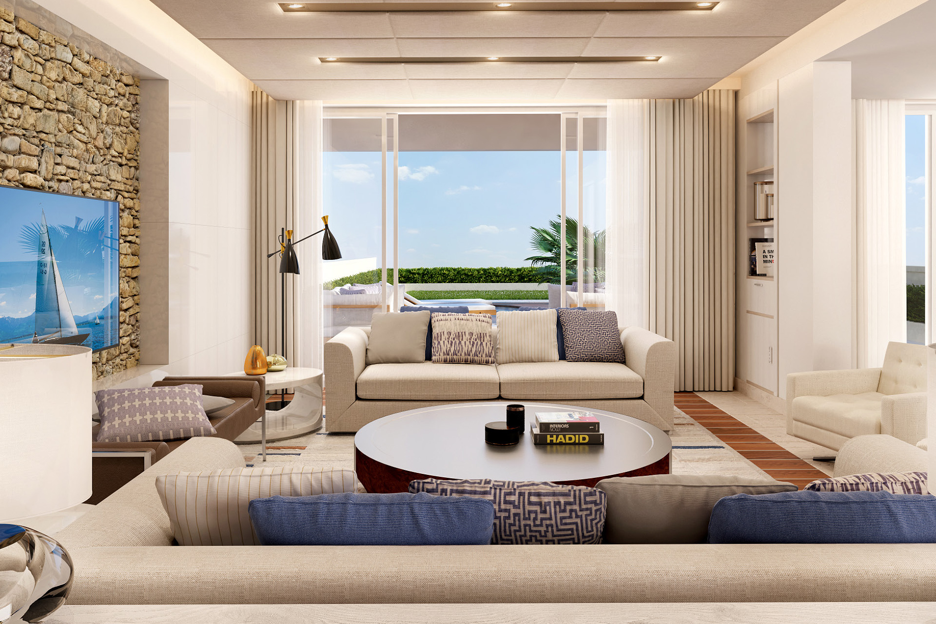 Modern living room with beige sofas and patterned pillows facing a round coffee table and large sliding glass doors opening to an outdoor seating area with a pool and greenery.