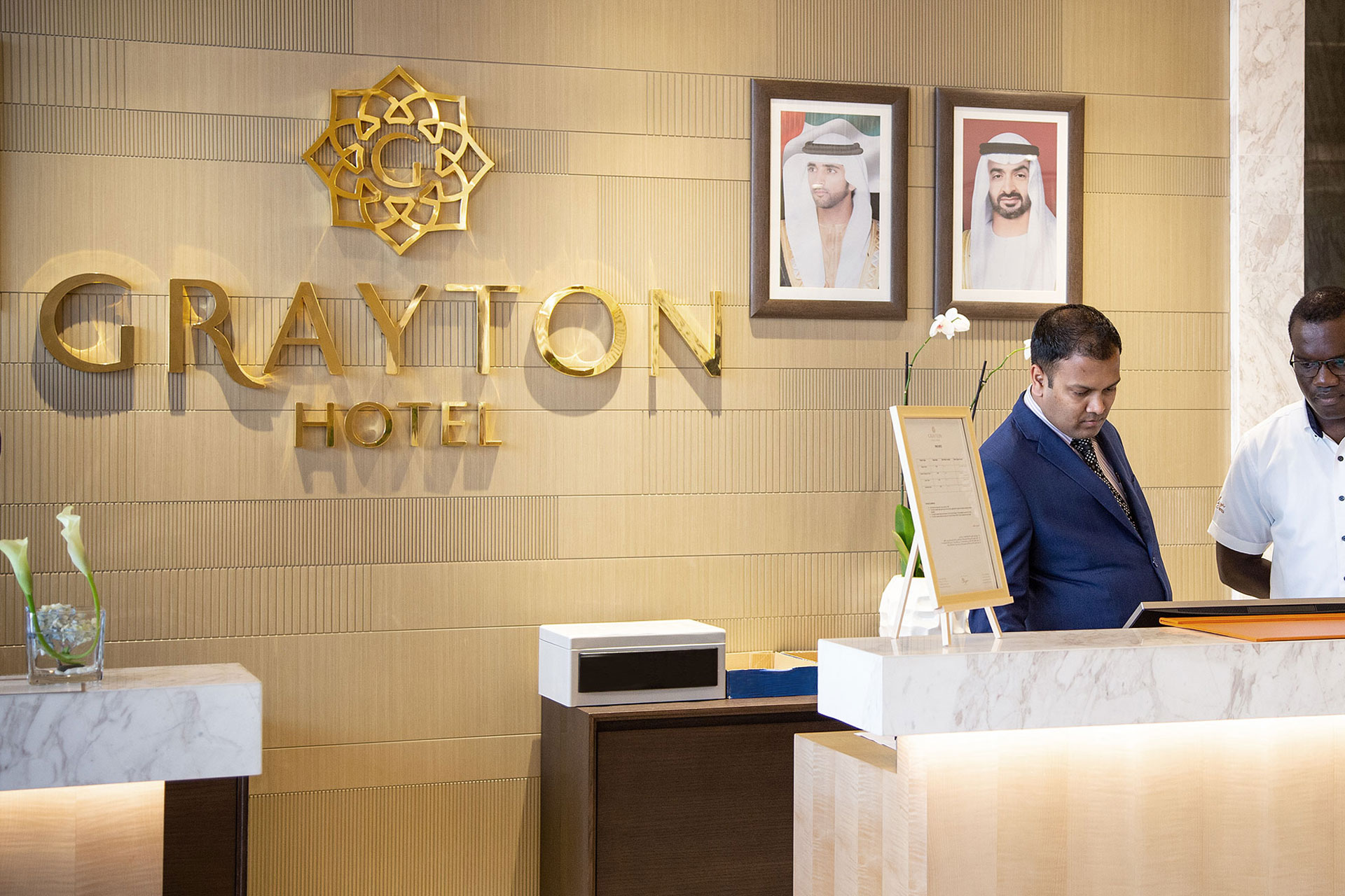 Reception area of Grayton Hotel with two staff members behind the marble counter and framed portraits on the wall.