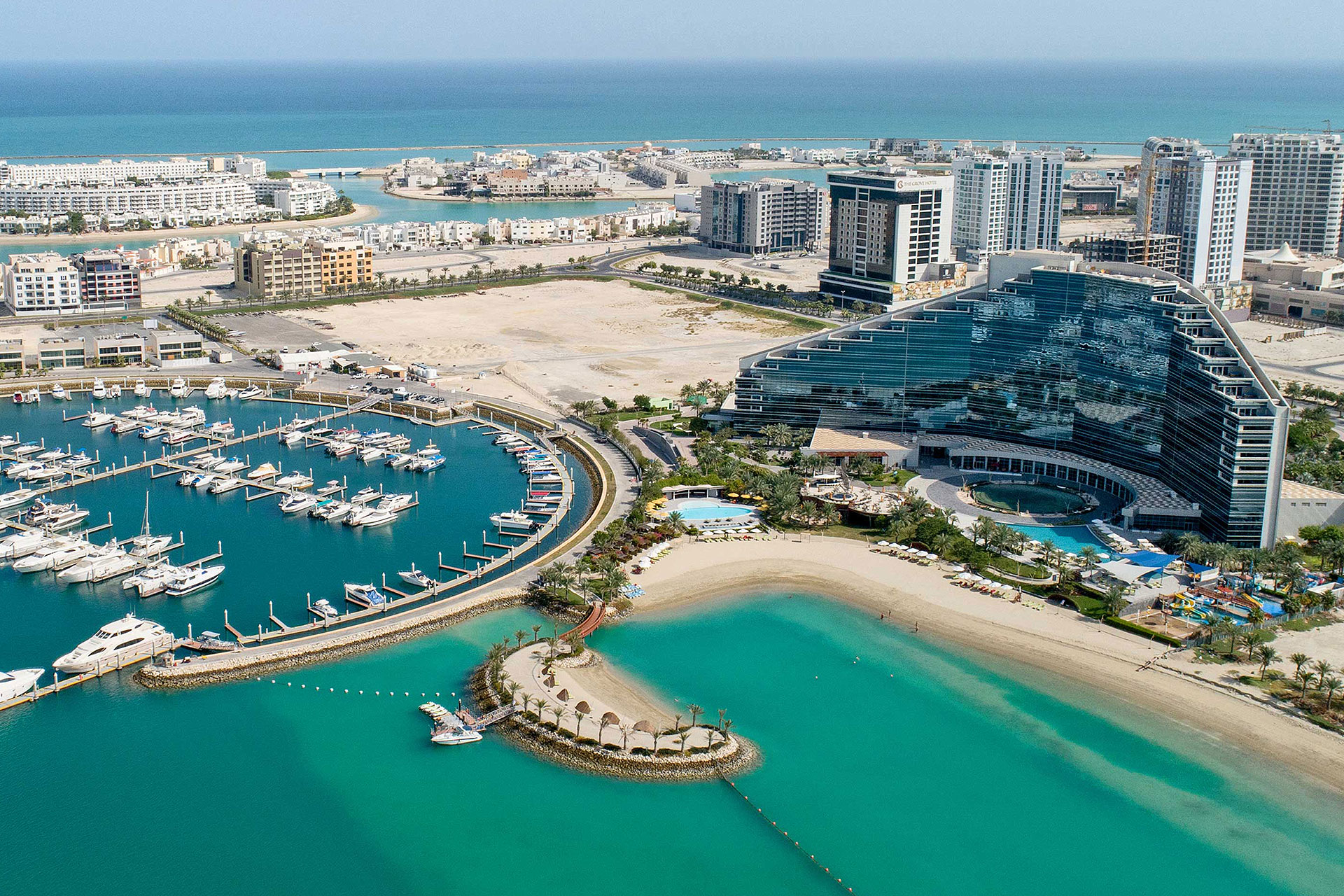 Aerial view of a curved luxury hotel beside a marina filled with yachts and turquoise waters with a sandy beach and surrounding buildings.
