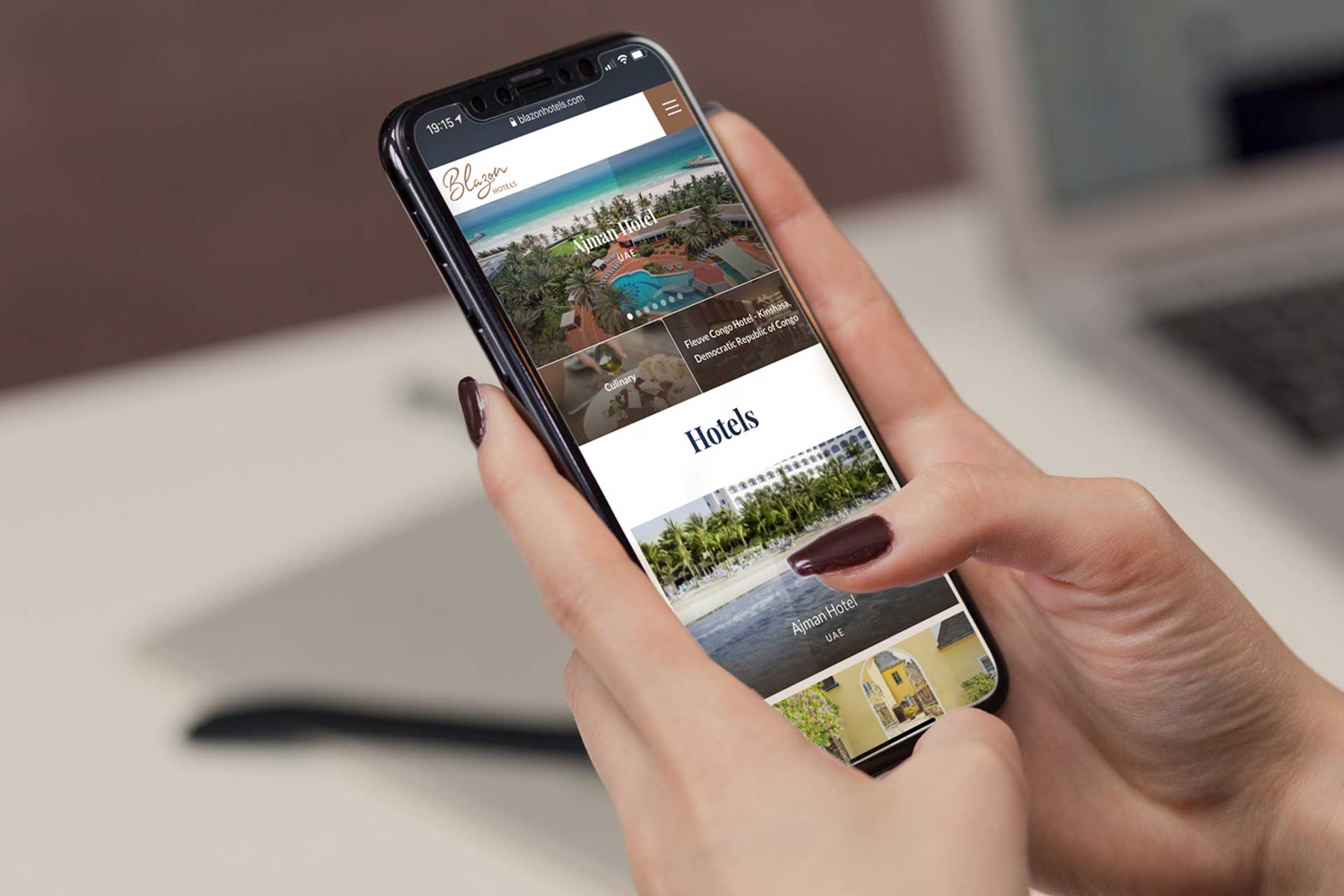 Person holding a smartphone displaying Blazon Hotels website featuring Ajman Hotel and other hotel listings.