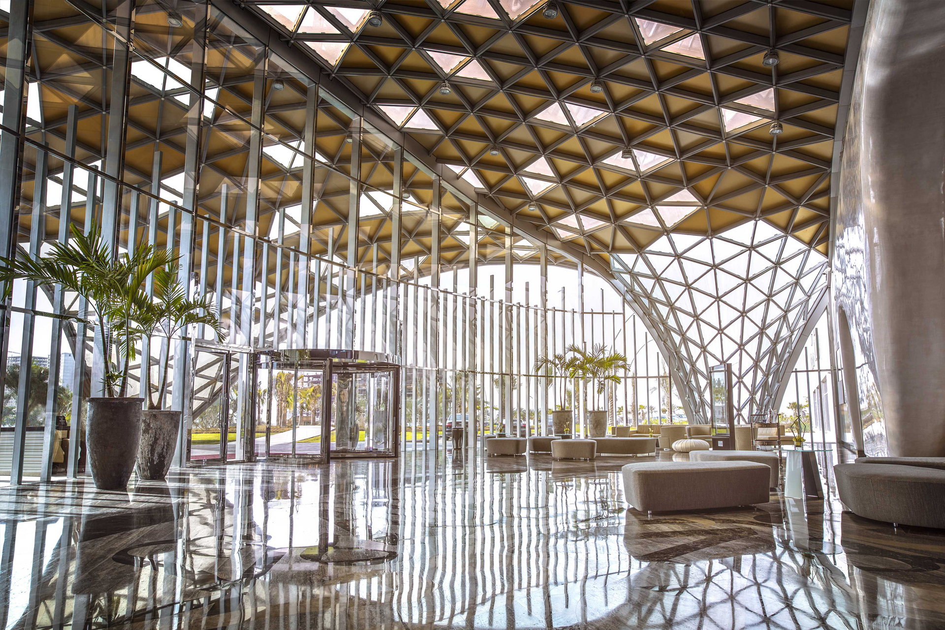 Spacious modern hotel lobby with high geometric glass ceiling, marble floor, potted plants, and minimalist seating.