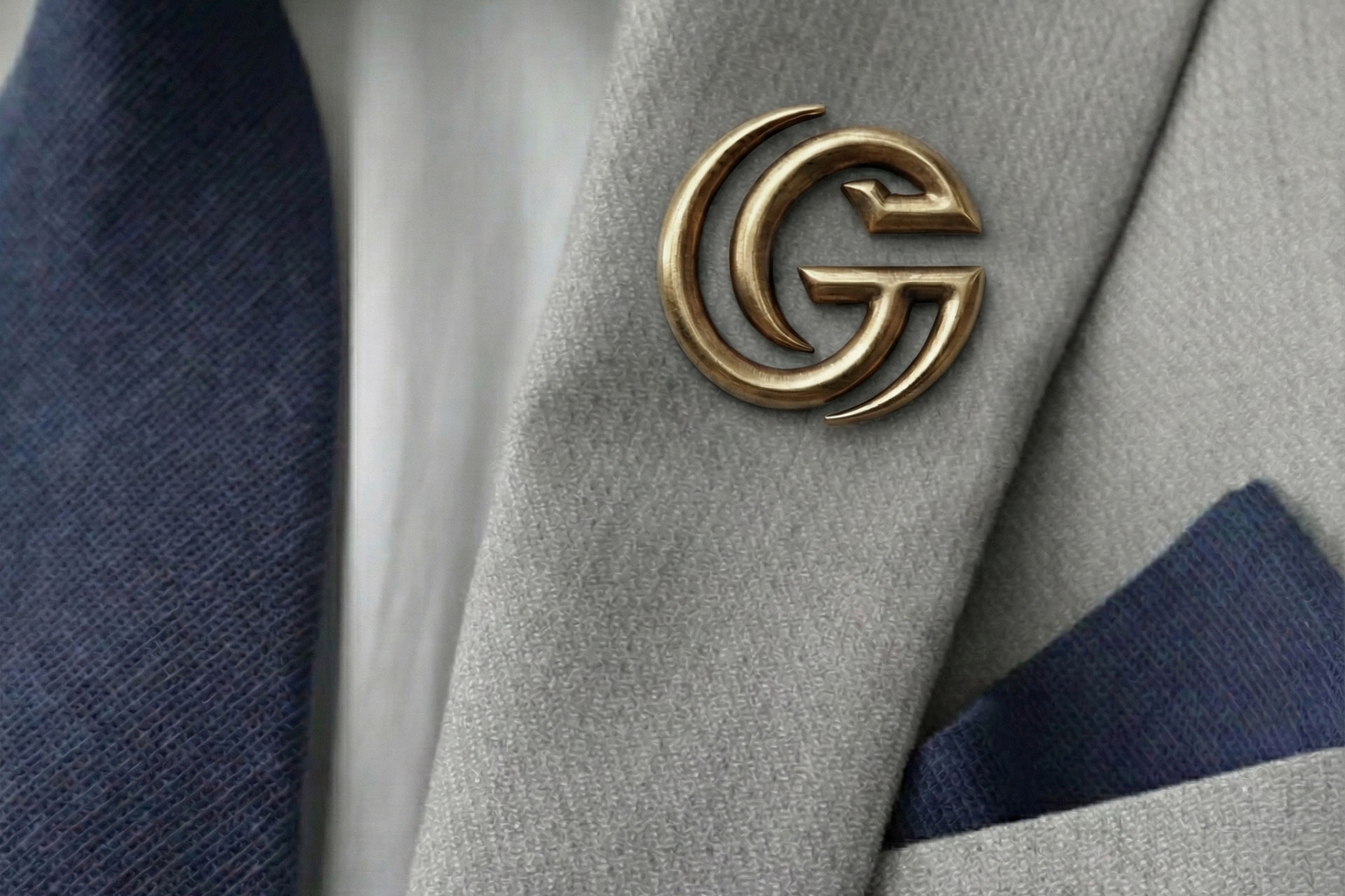 Gold GG logo pin on gray blazer lapel with navy blue pocket square and white shirt underneath.