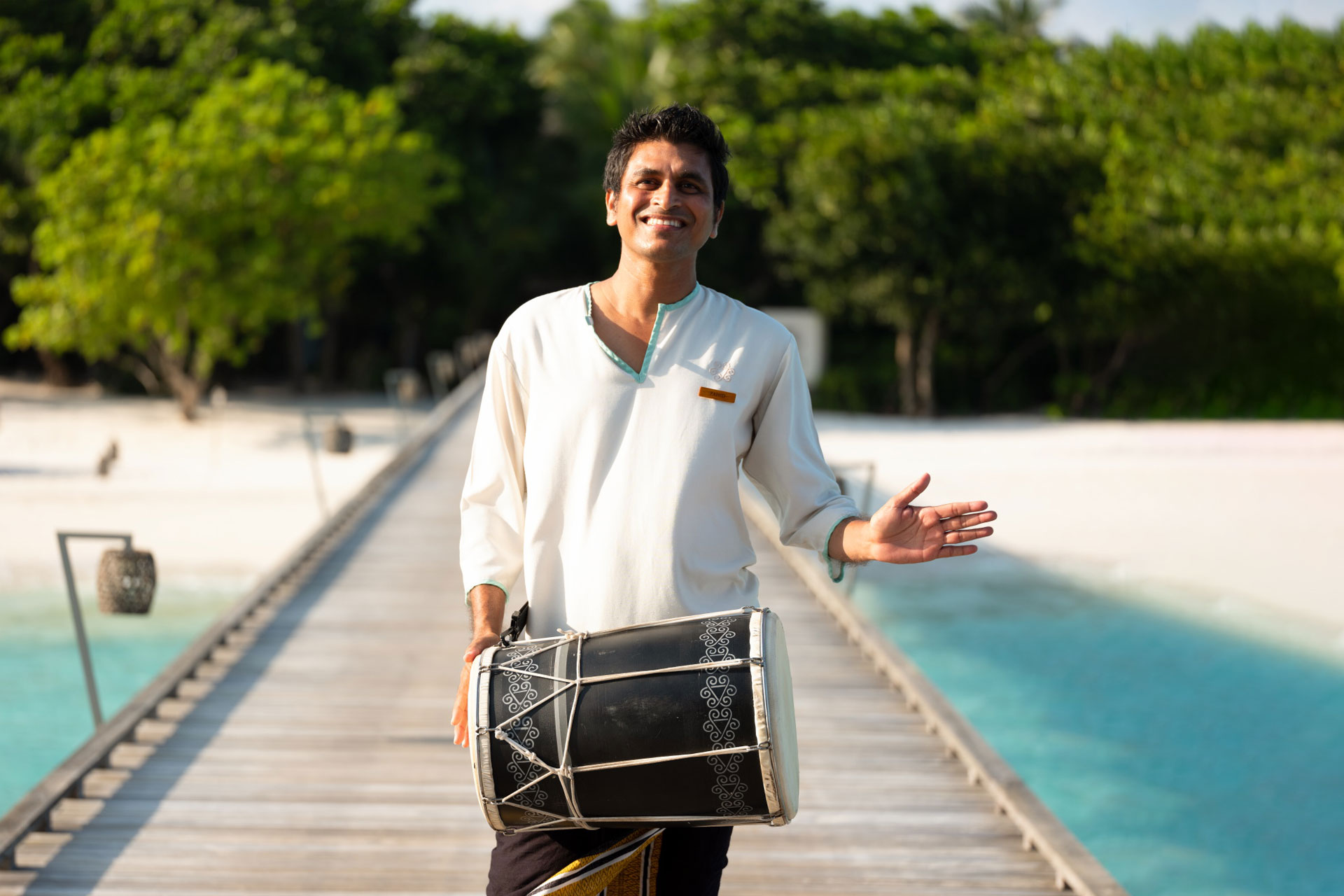 Smiling man in traditional attire playing a drum on a wooden pier by turquoise water and white sandy beach.