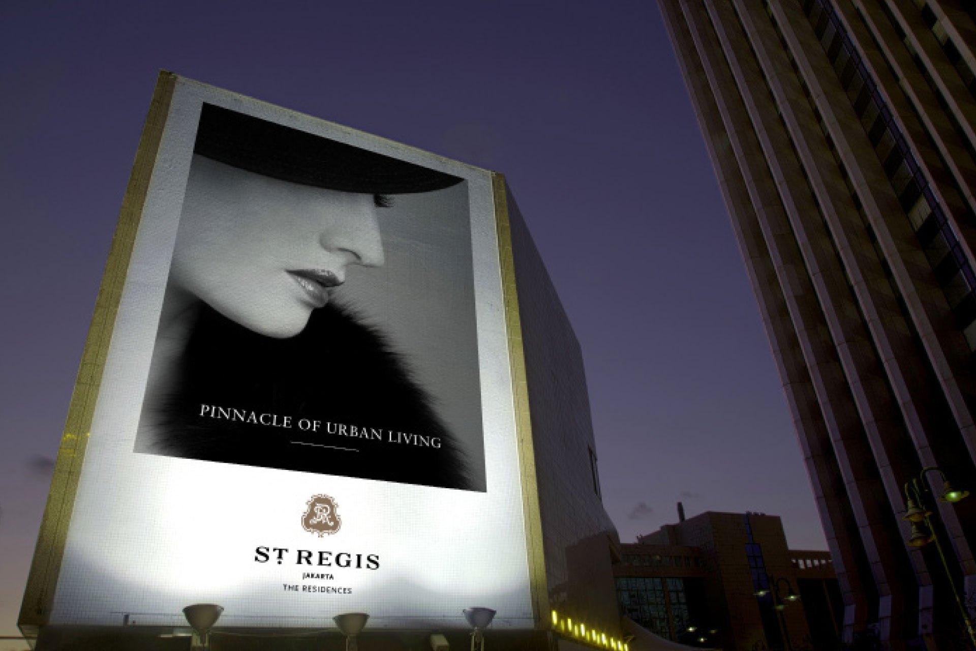 Brandteliers Singapore is a Luxury Hospitality branded residence Branding agency and a photograph of a Large illuminated billboard at dusk featuring a black-and-white close-up of a woman's face in profile wearing a hat and fur with text 'Pinnacle of Urban Living' and 'St Regis Jakarta The Residences'.