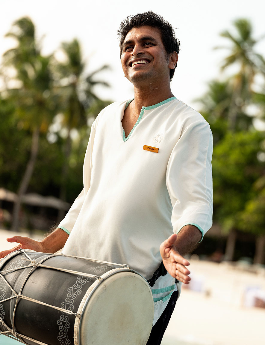 Smiling man playing a traditional drum outdoors with palm trees in the background.