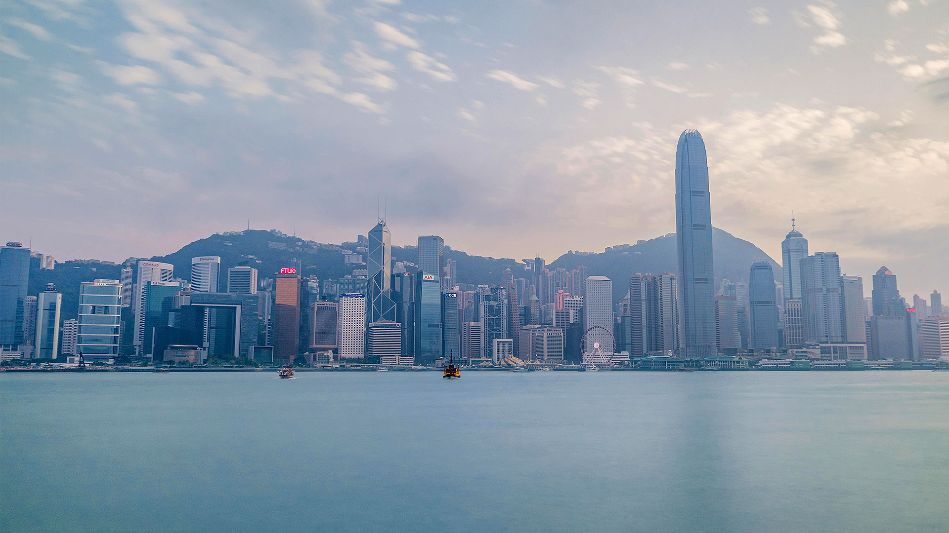Panoramic view of Hong Kong skyline with tall skyscrapers, Victoria Harbour, and mountainous background under a cloudy sky.