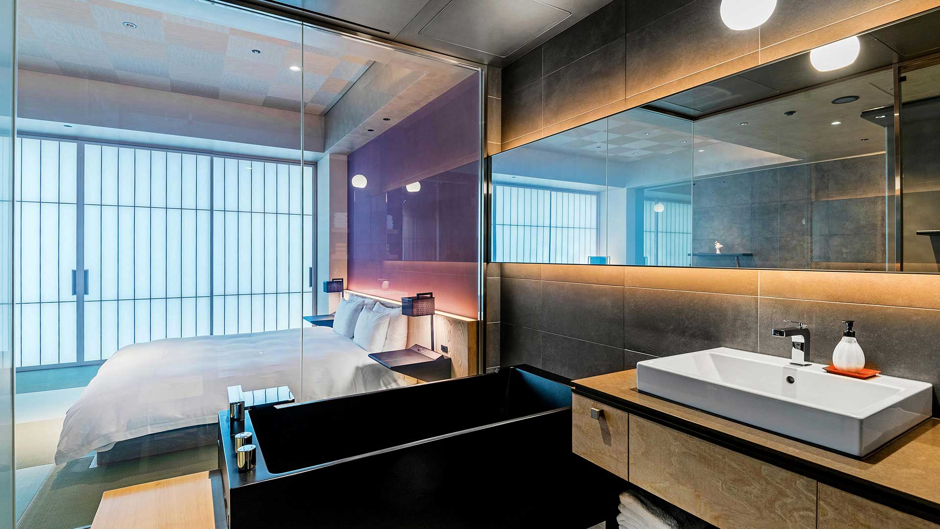Modern hotel room with a white bed behind glass partition and a black bathtub with a large mirror and sink in the foreground.
