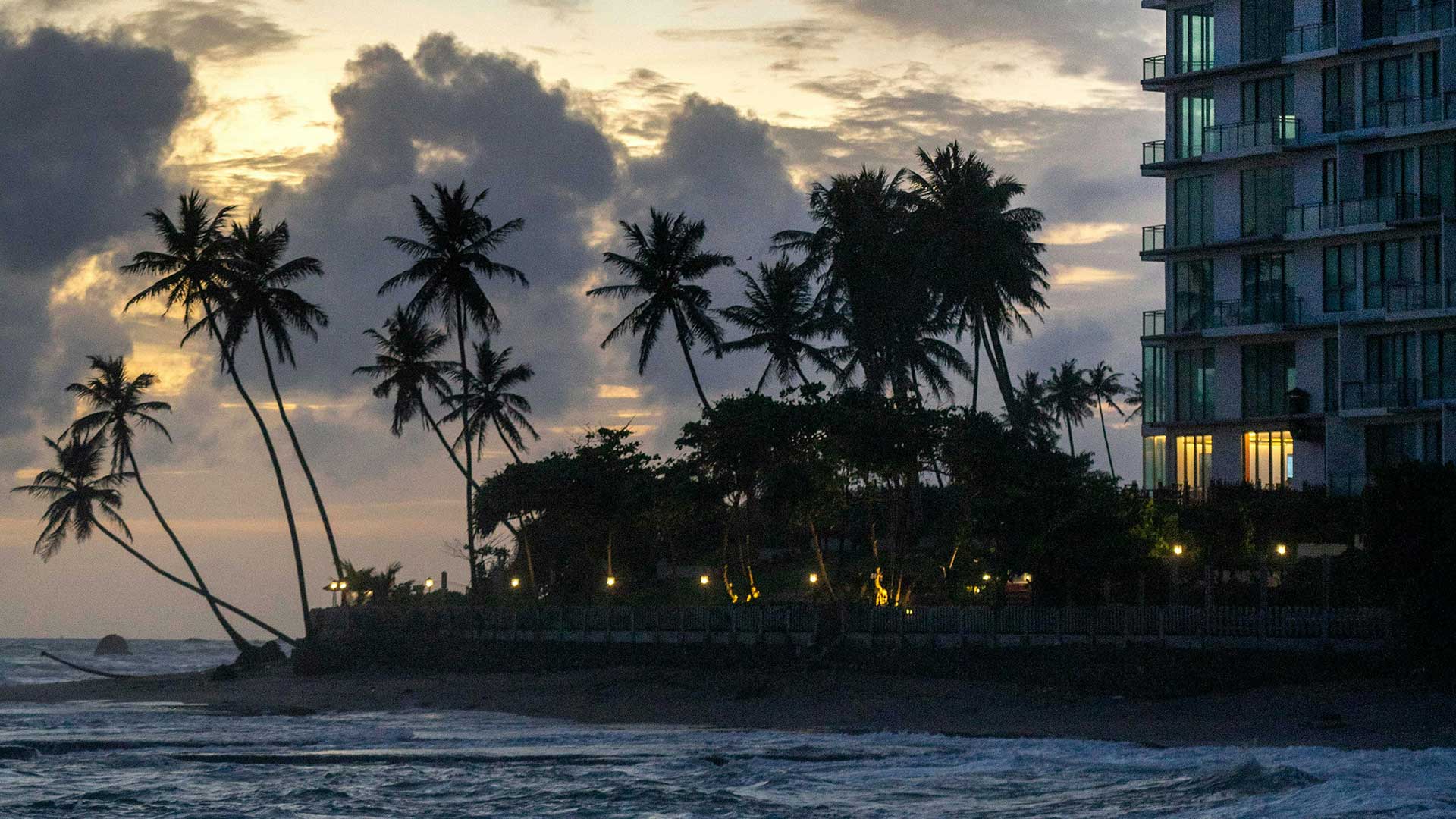 Palm trees in Sri Lanka silhouetted against a cloudy sunset sky near the Indian ocean with a modern building on the right side.