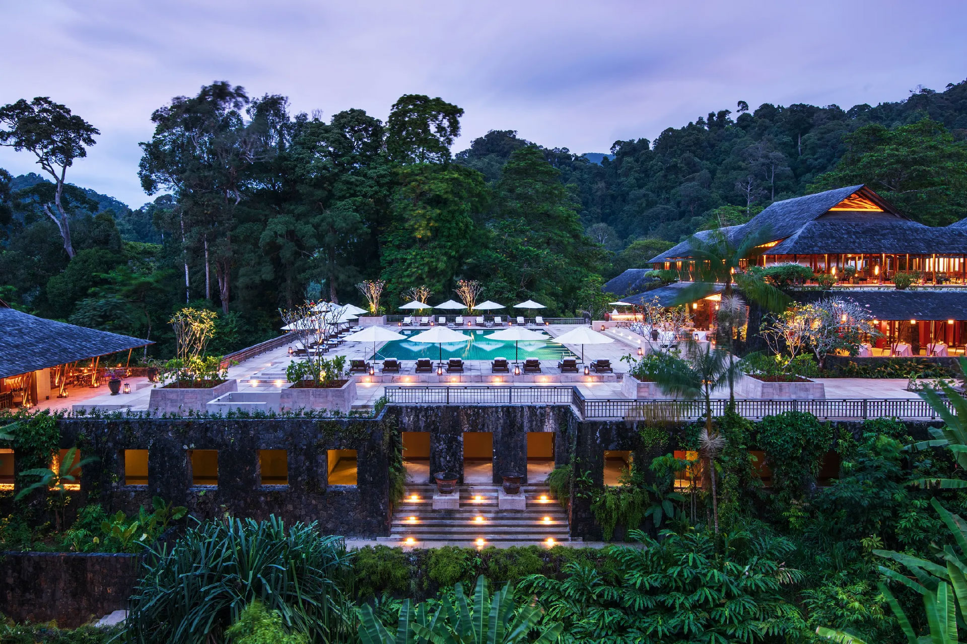 Luxurious resort with an illuminated pool surrounded by lounge chairs and umbrellas amidst lush forest at dusk.