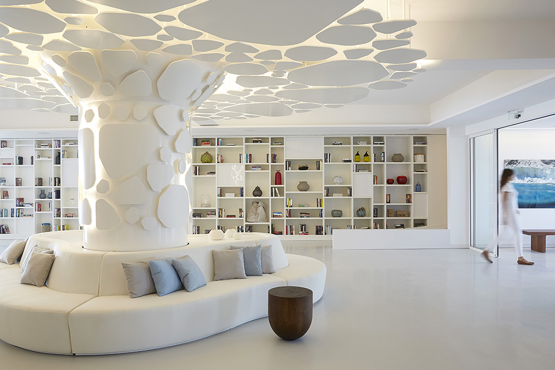 Modern white lounge area with curved seating around a sculpted pillar and built-in shelves filled with books and decor, a person walking near a glass door.
