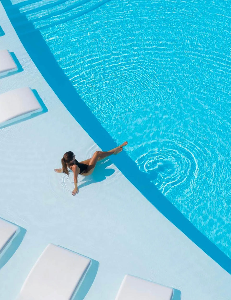 Woman in a black swimsuit sitting at the edge of a blue swimming pool surrounded by white lounge chairs.