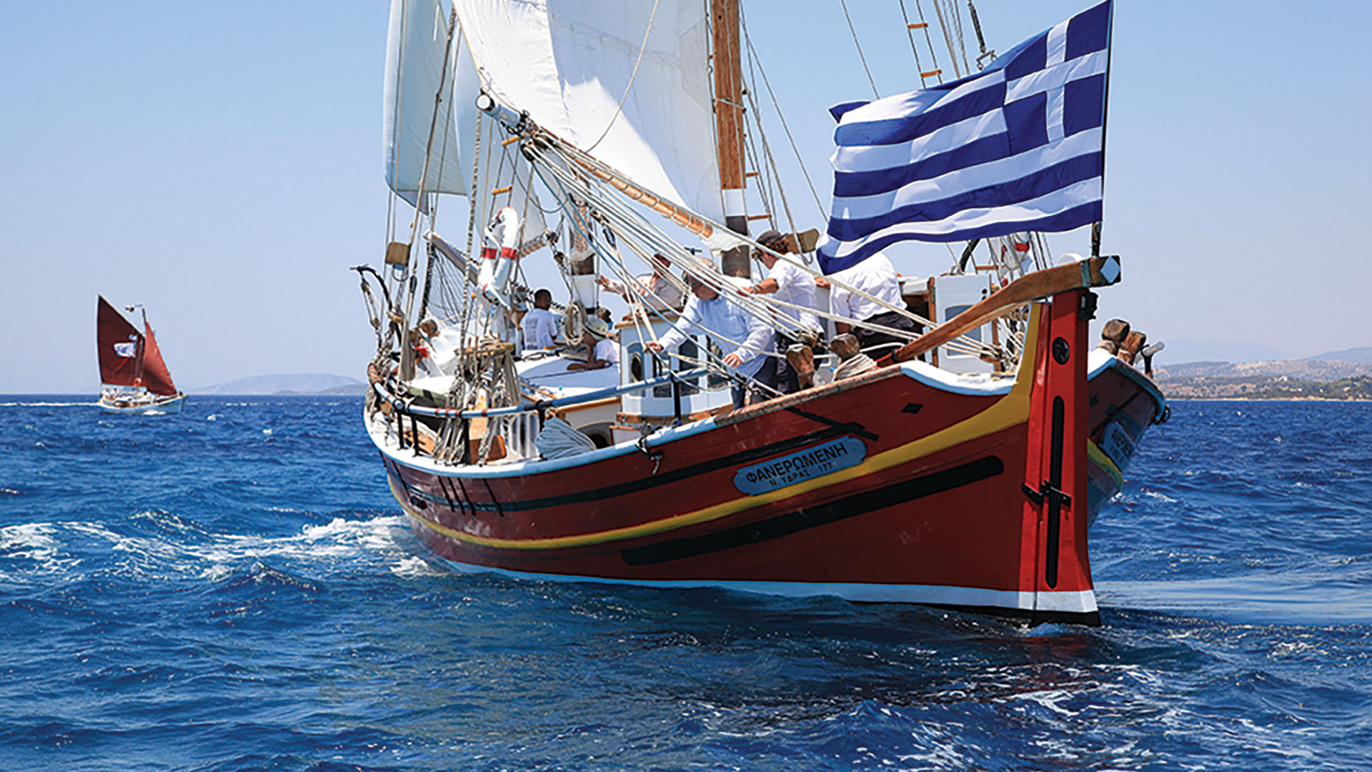 Sailing boat with white sails and a Greek flag on blue sea with people on board on a sunny day.