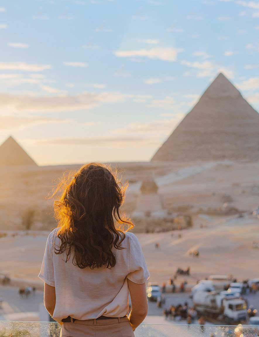 Woman with curly hair standing and looking at the pyramids of Giza at sunset.
