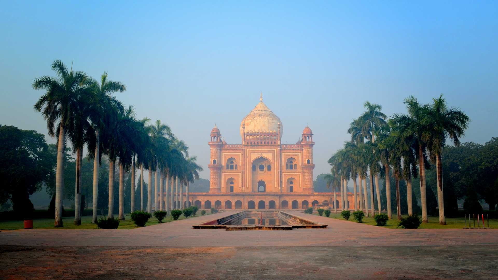 Symmetrical view of a historic sandstone mausoleum with a white dome, flanked by rows of tall palm trees and a reflecting pool in front.