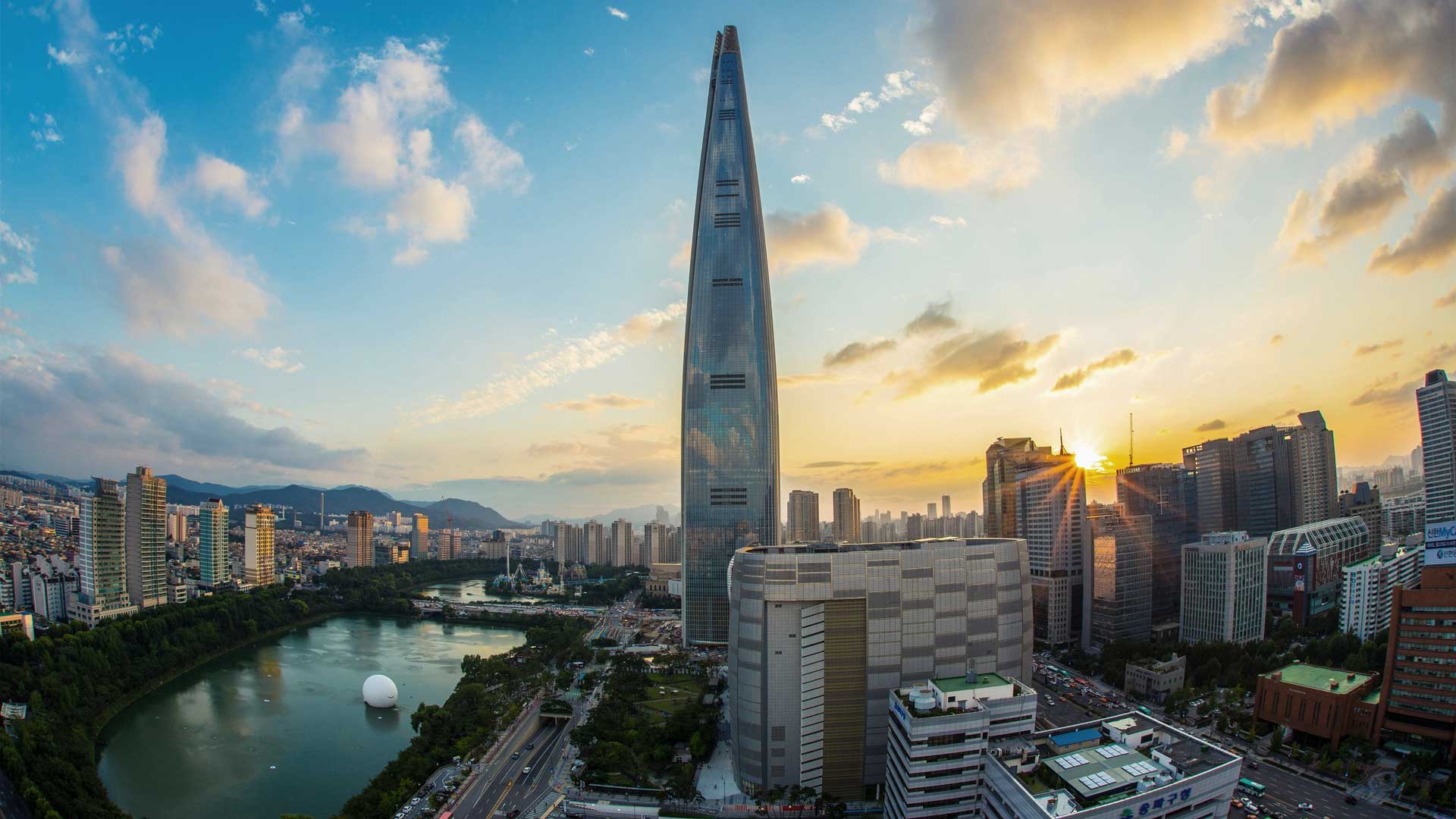 Cityscape of Seoul at sunset featuring the tall, sleek Lotte World Tower beside a winding river and surrounding skyscrapers.