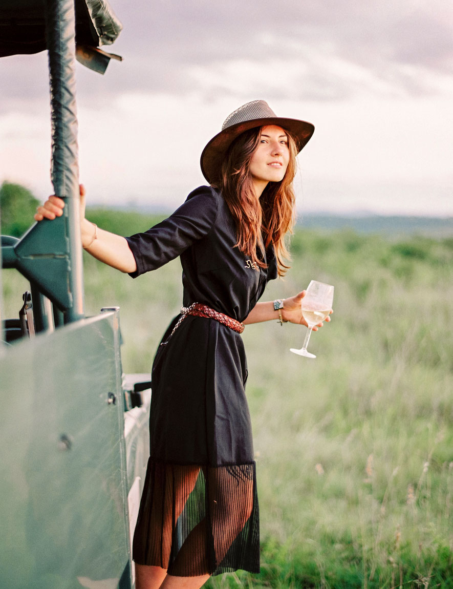 Woman in a black dress and hat holding a glass of white wine standing by a safari vehicle in a grassy field.