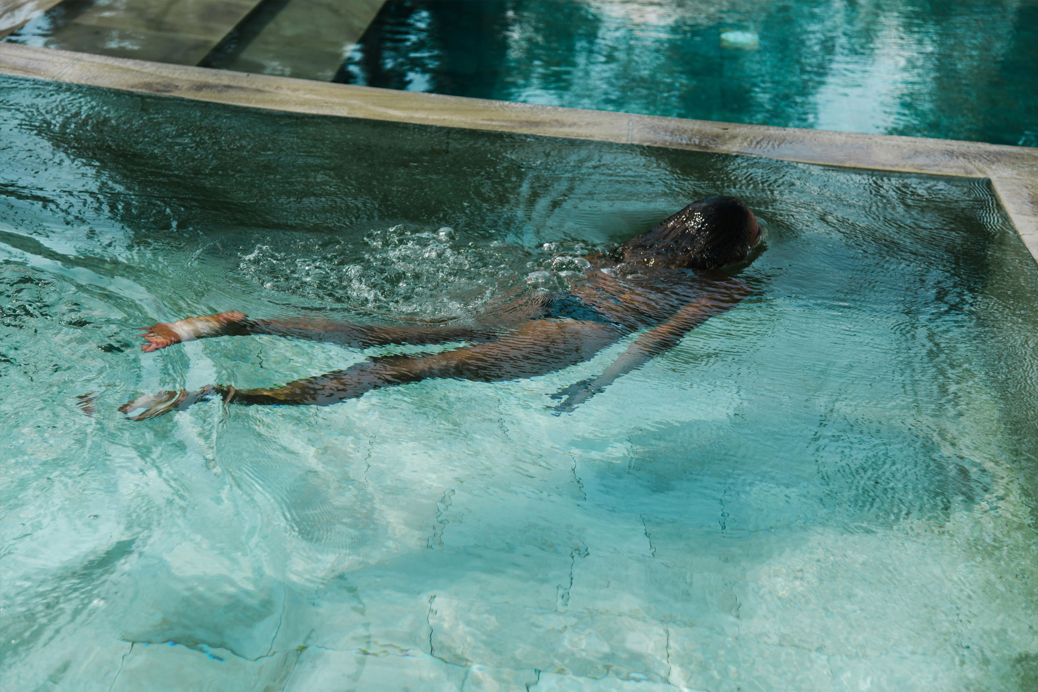 Person floating face down in a clear blue swimming pool near the edge.