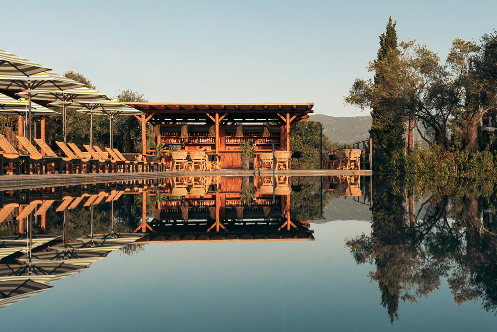 Wooden poolside bar and lounge chairs with umbrellas reflected in a calm swimming pool.