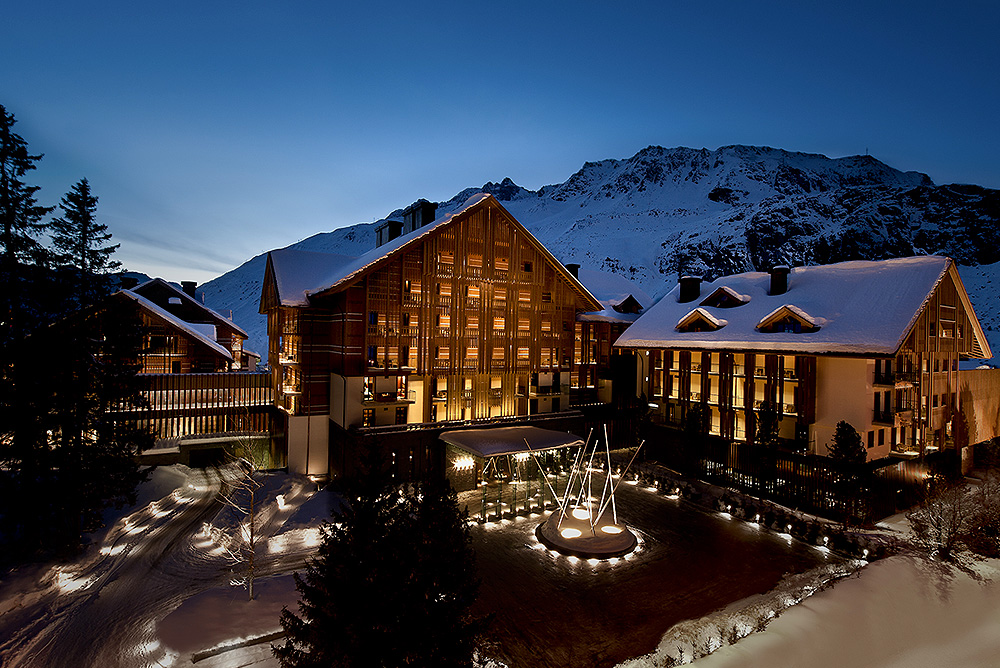 Snow-covered alpine hotel buildings lit warmly at dusk with mountains in the background.