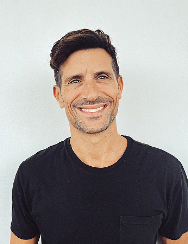 Smiling man with short dark hair and light stubble wearing a black t-shirt against a white background.