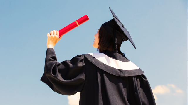 Graduate holding up a red diploma against a clear blue sky