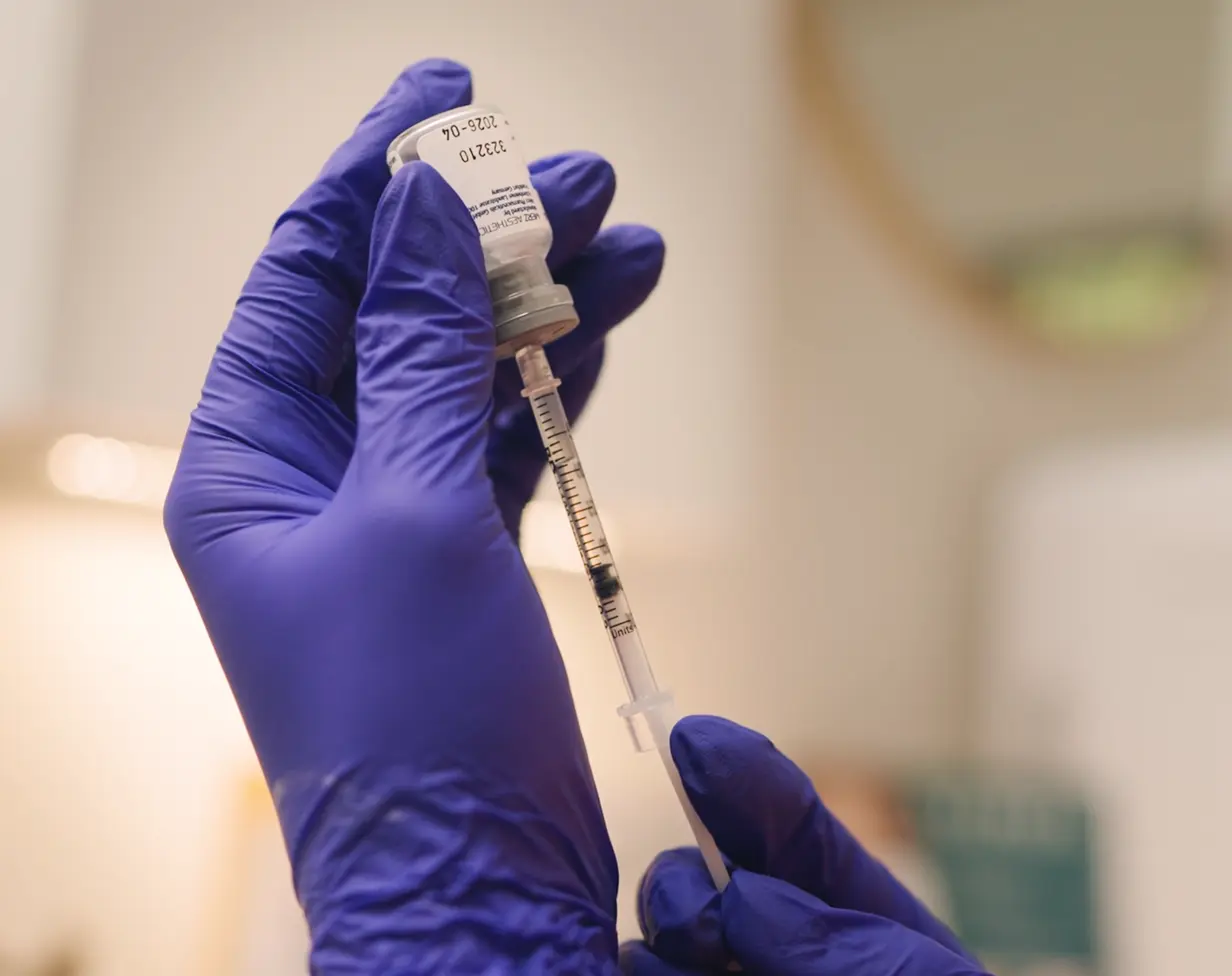 A gloved hand holding a vial while drawing liquid into a syringe for an injection.