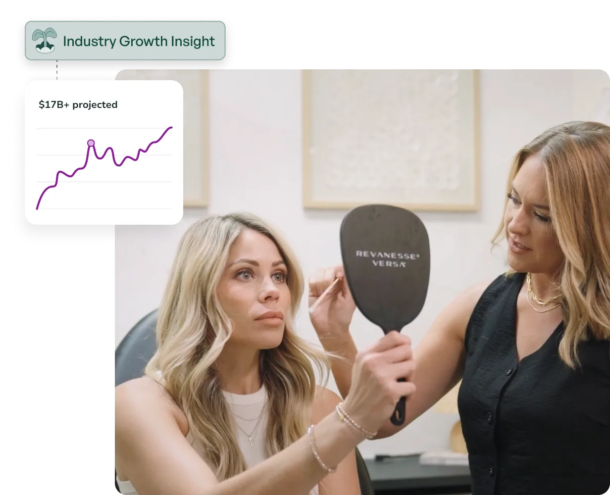 A woman holding a Revansese Versa hand mirror looks at her reflection while another woman assists her in a clinical or spa setting.