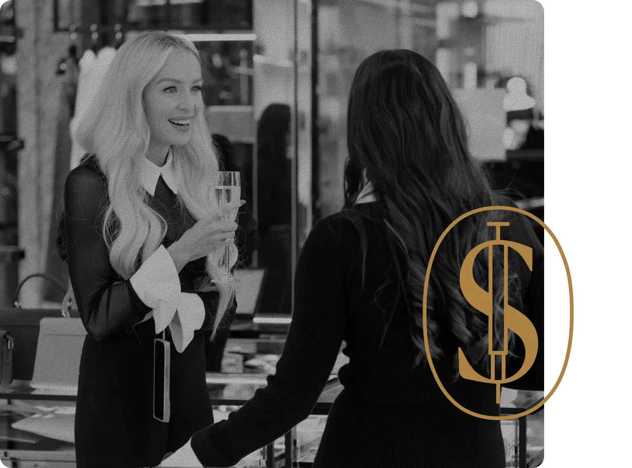 Two women conversing indoors, one holding a champagne glass and smiling, with the other seen from behind.