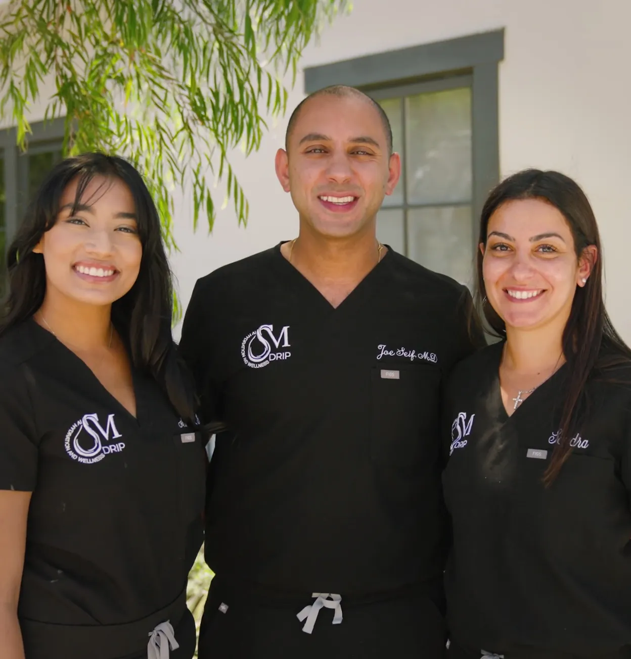 Three smiling healthcare professionals in black scrubs standing outside in front of a building with windows and greenery.