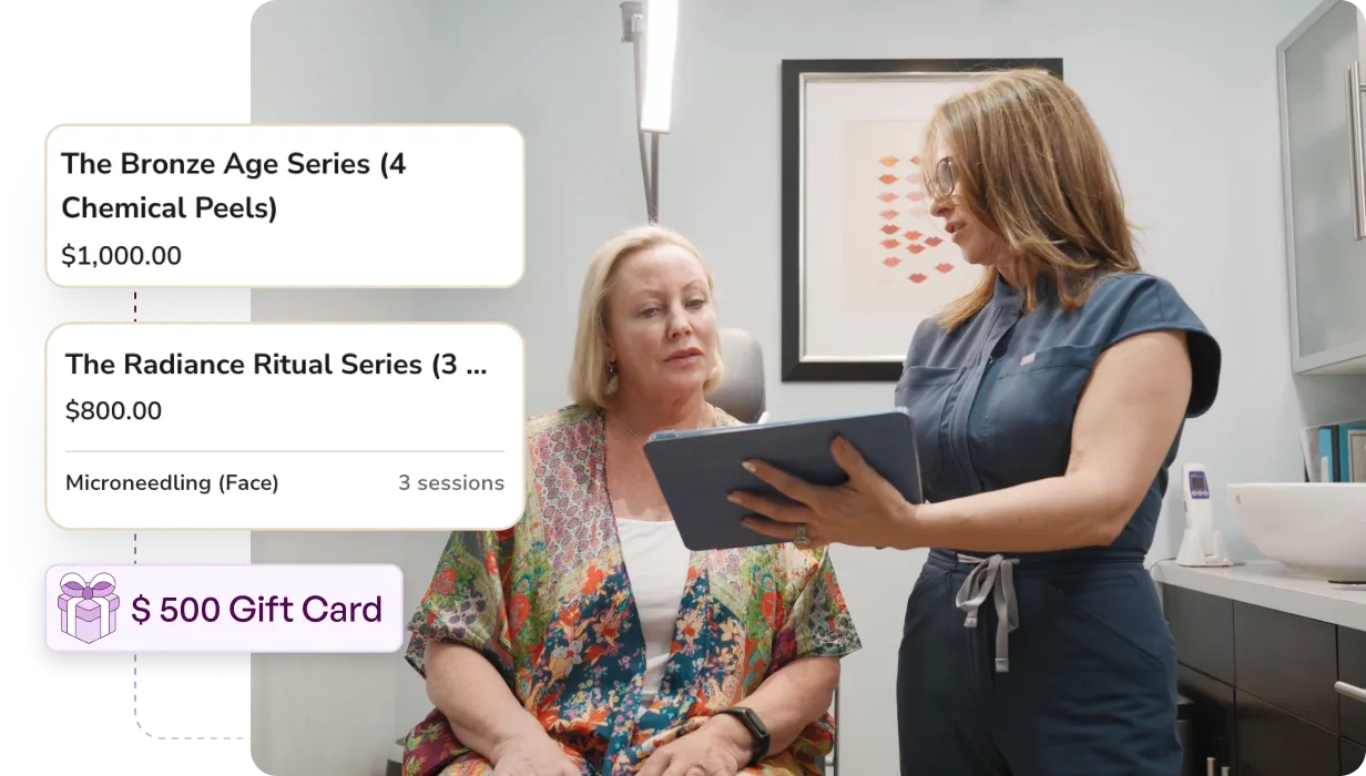 A healthcare professional in blue scrubs showing a tablet to a seated woman in a floral robe inside a medical office.