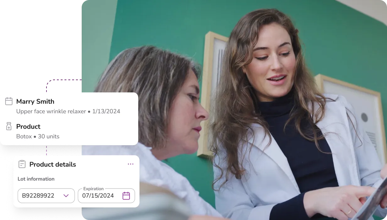 Two women looking at a tablet; one in a white coat appears to be explaining something to the other, with green wall and framed pictures in the background.