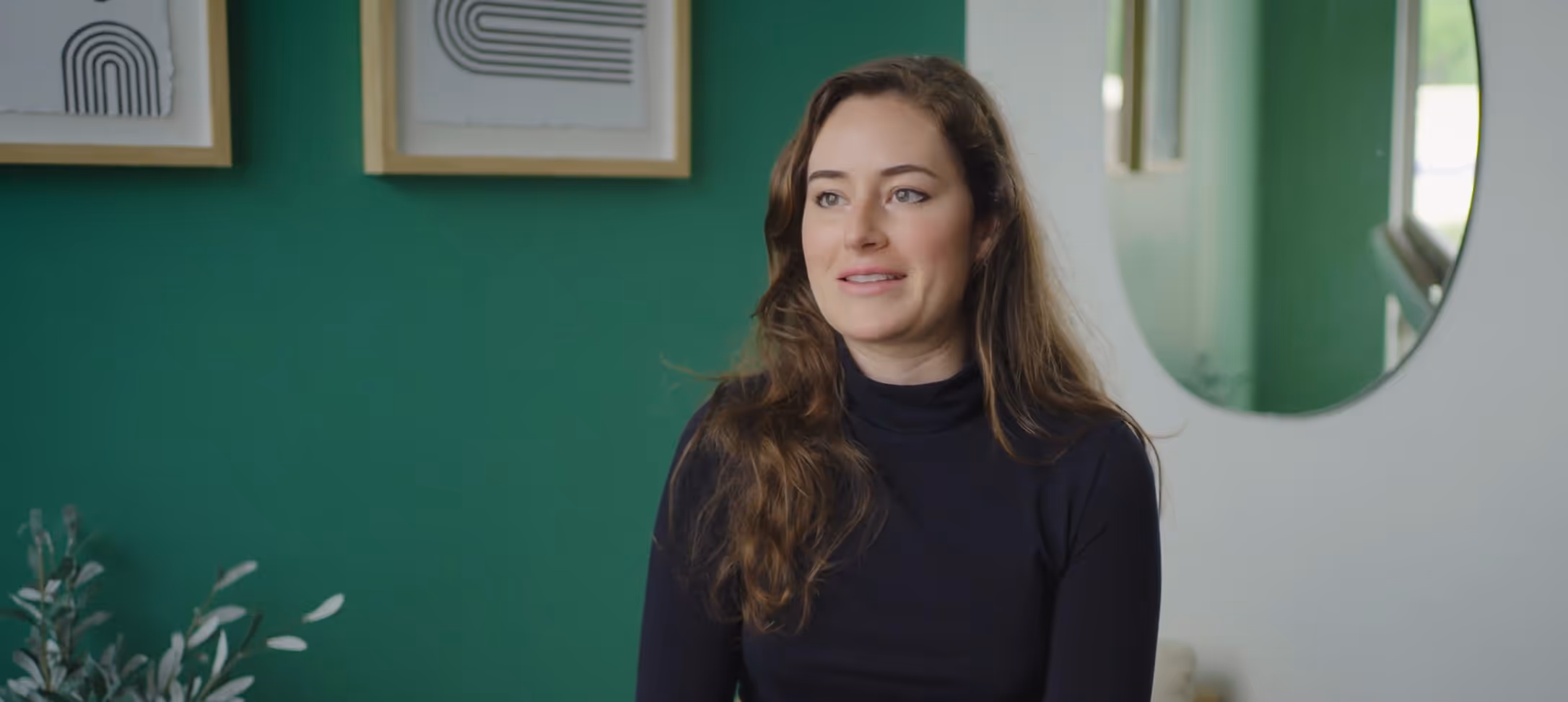Woman with long brown hair in a black turtleneck sitting in front of a green wall with framed abstract art and a round mirror.