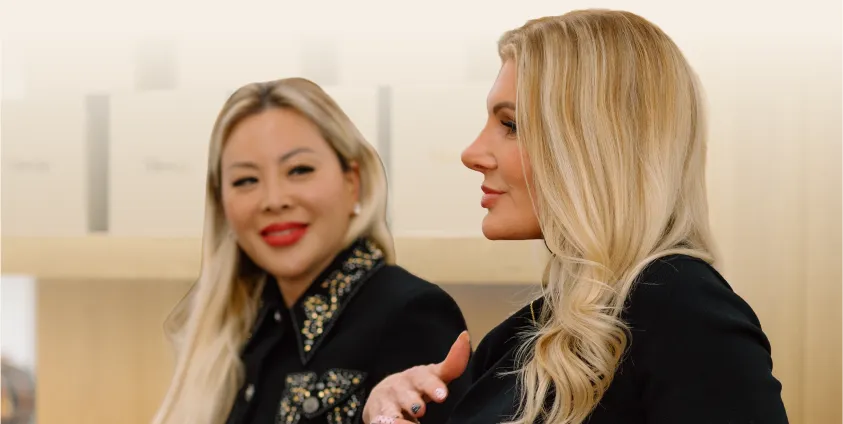 Two professionally dressed women smiling and engaged in conversation at a Moxie event, with branded product packaging displayed on shelves in the background