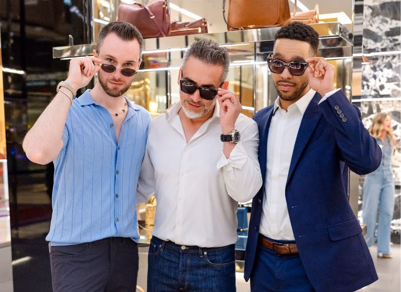 Three men in front of a handbag display, each adjusting their sunglasses.
