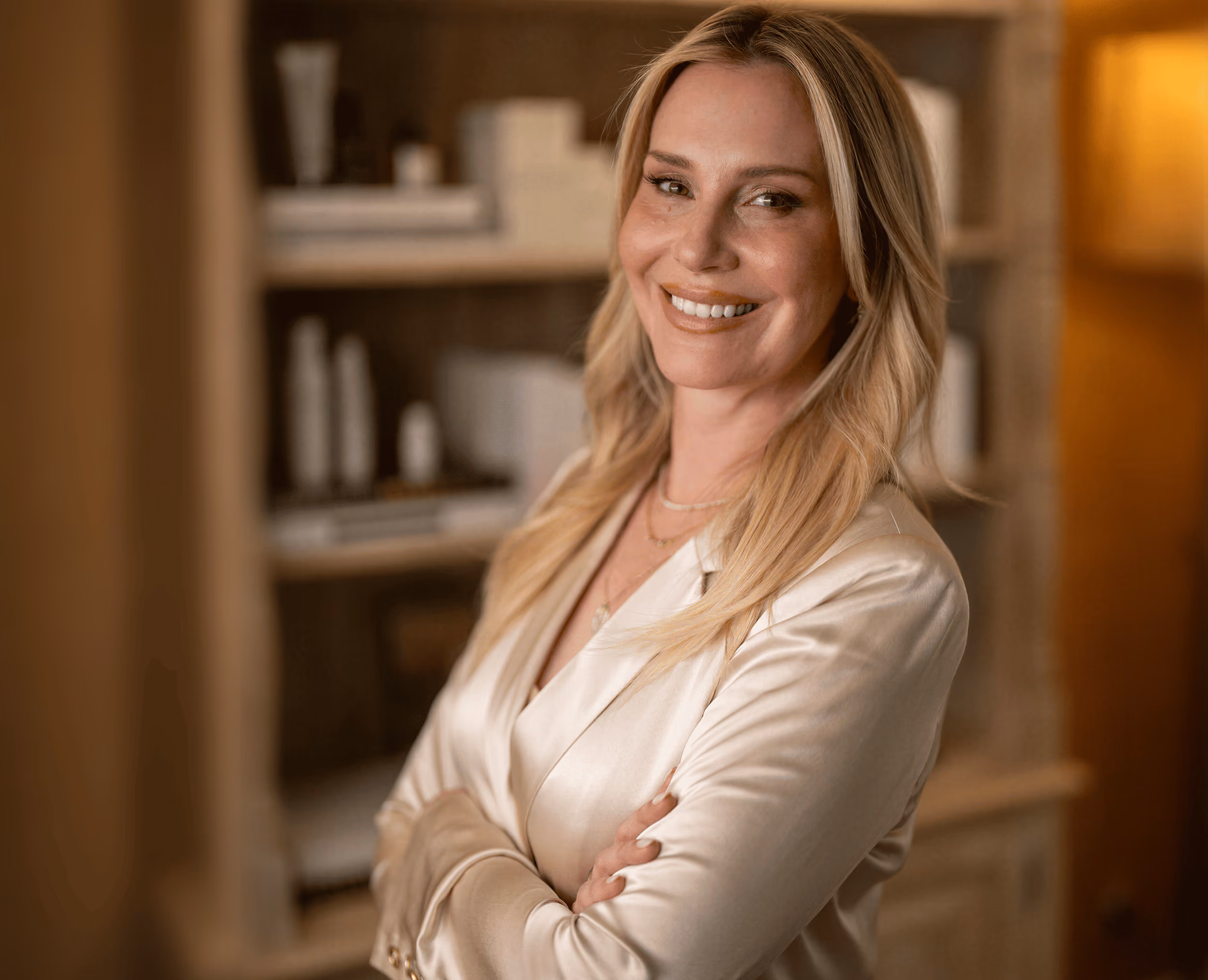 Smiling blonde woman with arms crossed, wearing a cream satin blazer, standing in front of a softly lit bookshelf.