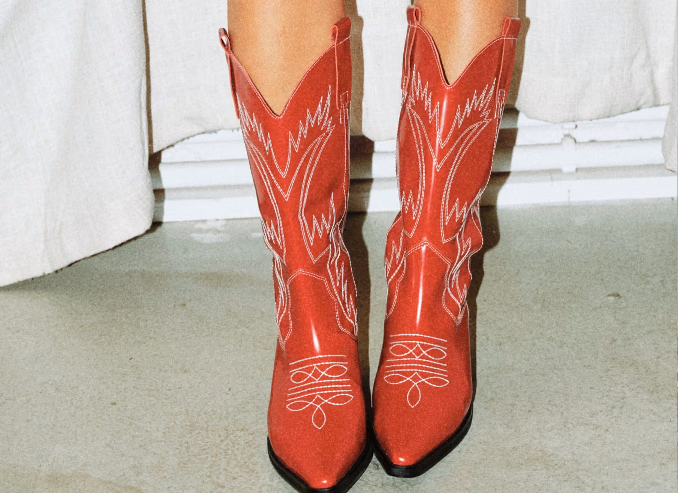 Person wearing tall red cowboy boots with white western stitching and pointed toes, standing on a pale concrete floor before a white curtain.