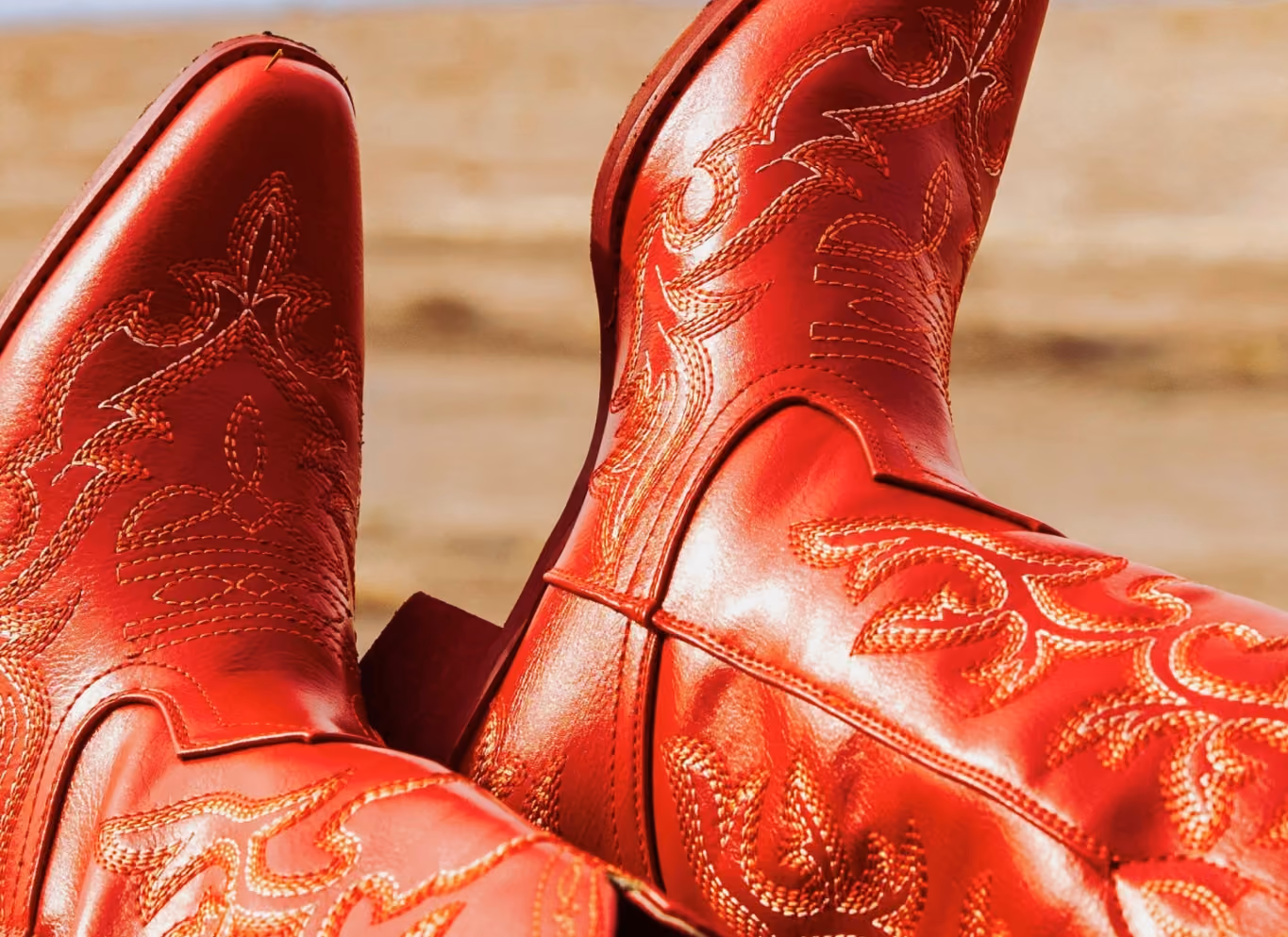 Close-up of glossy red leather cowboy boots with ornate tonal stitching, against a blurred sandy desert background.