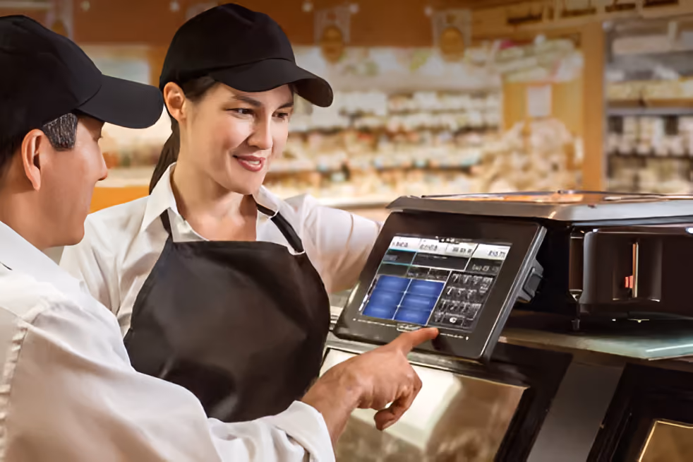 Two employees wearing black caps and aprons operating a digital touchscreen device in a supermarket or store setting.