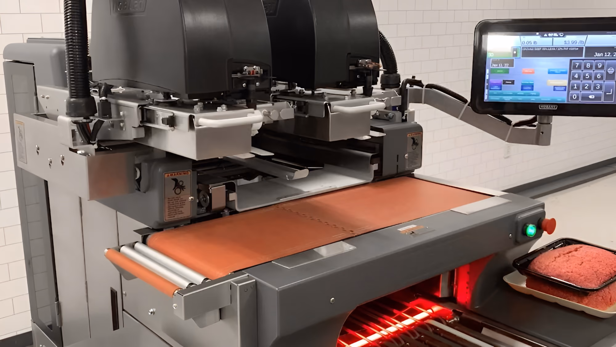 Industrial meat processing machine with dual heads and a conveyor belt, next to a digital control panel displaying operational data, with trays of ground meat visible nearby.
