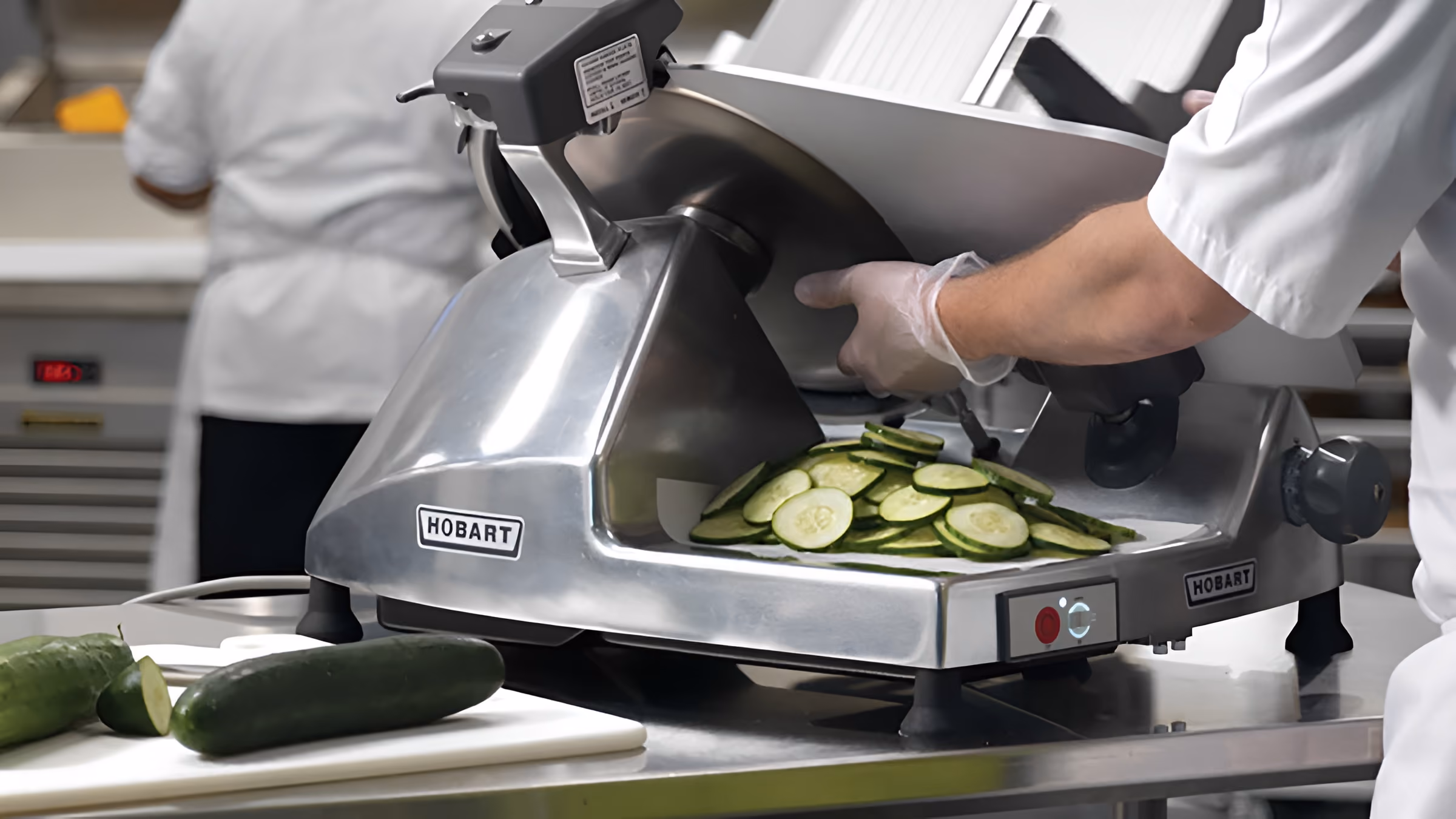Person using a stainless steel Hobart commercial slicer to cut cucumbers into thin slices on a kitchen counter with two whole cucumbers nearby on a cutting board.