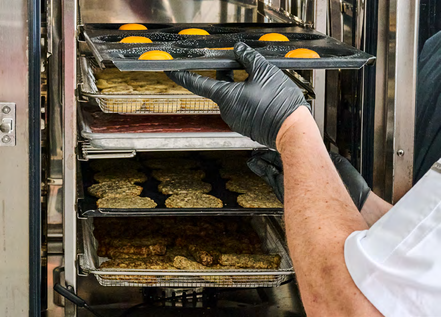 Person wearing black gloves placing a baking tray with round pastries topped with bright orange yolks into a commercial oven; several other trays with baked goods are inside the oven.