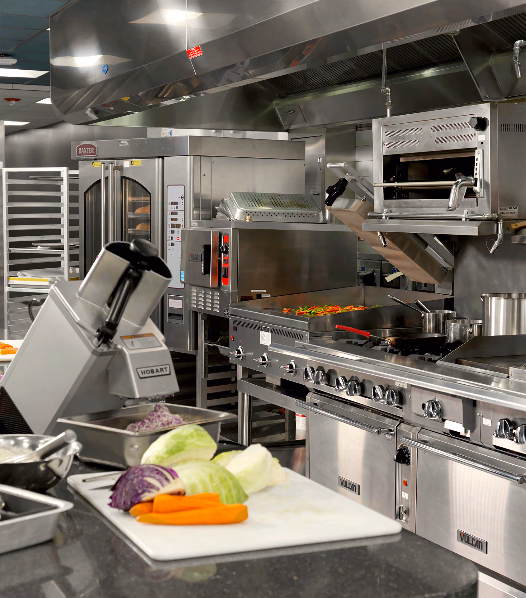 Commercial kitchen with stainless steel appliances including a Hobart slicer, a Vulcan stove with multiple burners, an oven, and preparation area with chopped vegetables including cabbage, carrots, and purple cabbage on a white cutting board.