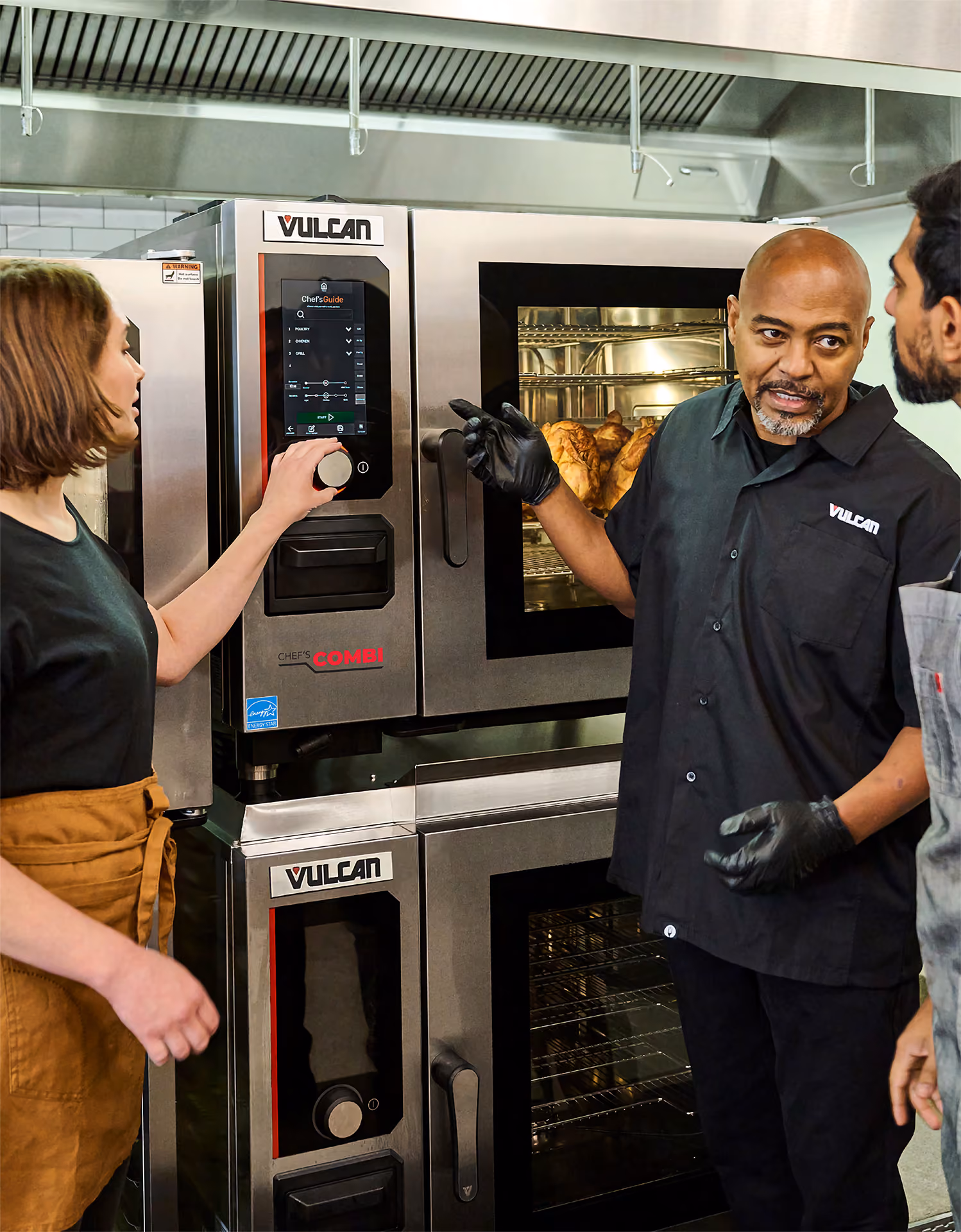 Three people in a professional kitchen are gathered around stainless steel Vulcan Chef's Combi ovens; one person is adjusting the oven controls while another explains or gestures towards the oven with cooked food visible inside.