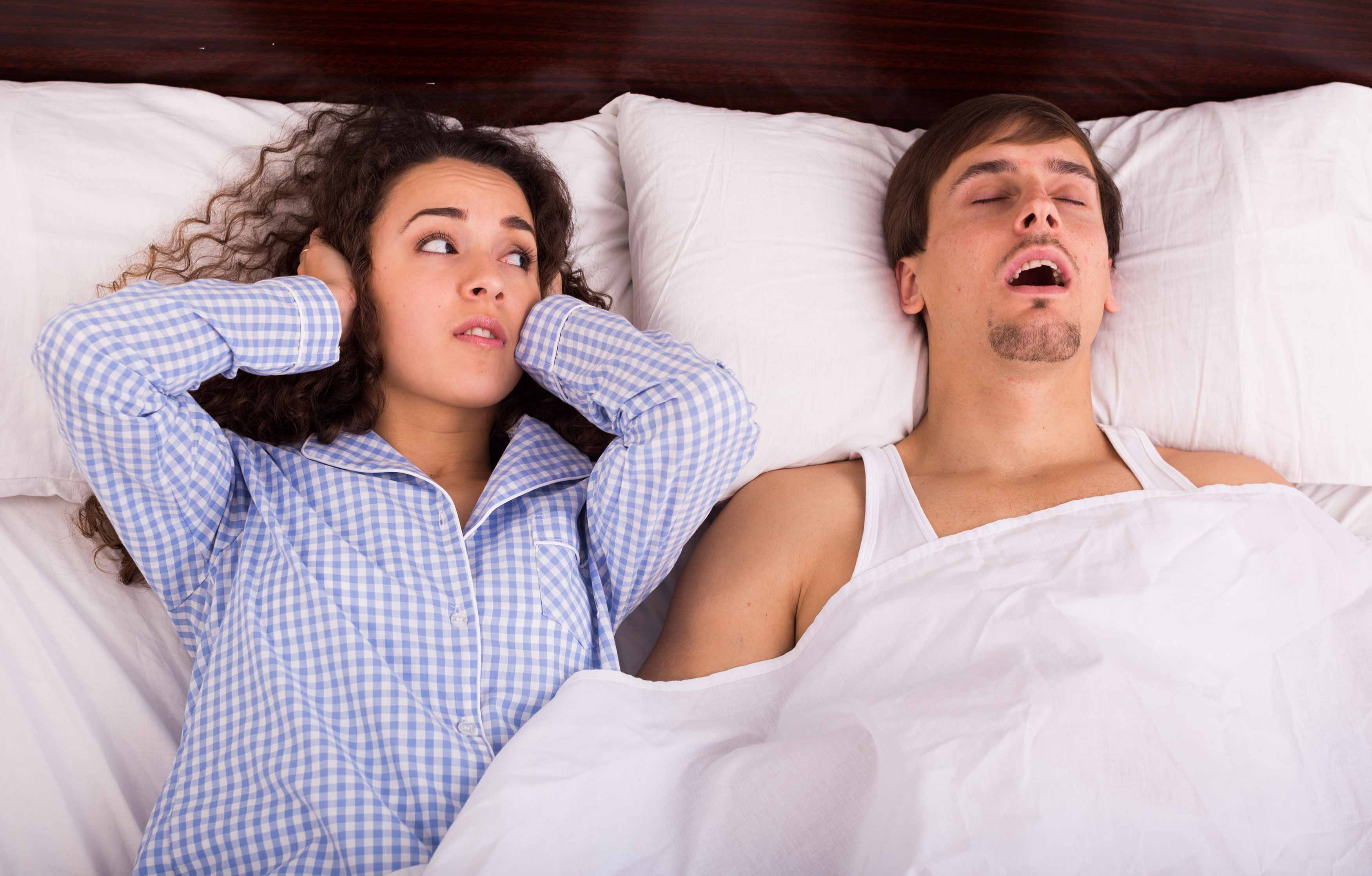 This has a couple in bed sleeping. The male is snoring and keeping his partner asleep. She is looking at him annoyed with her hands covering her ears because of his snoring