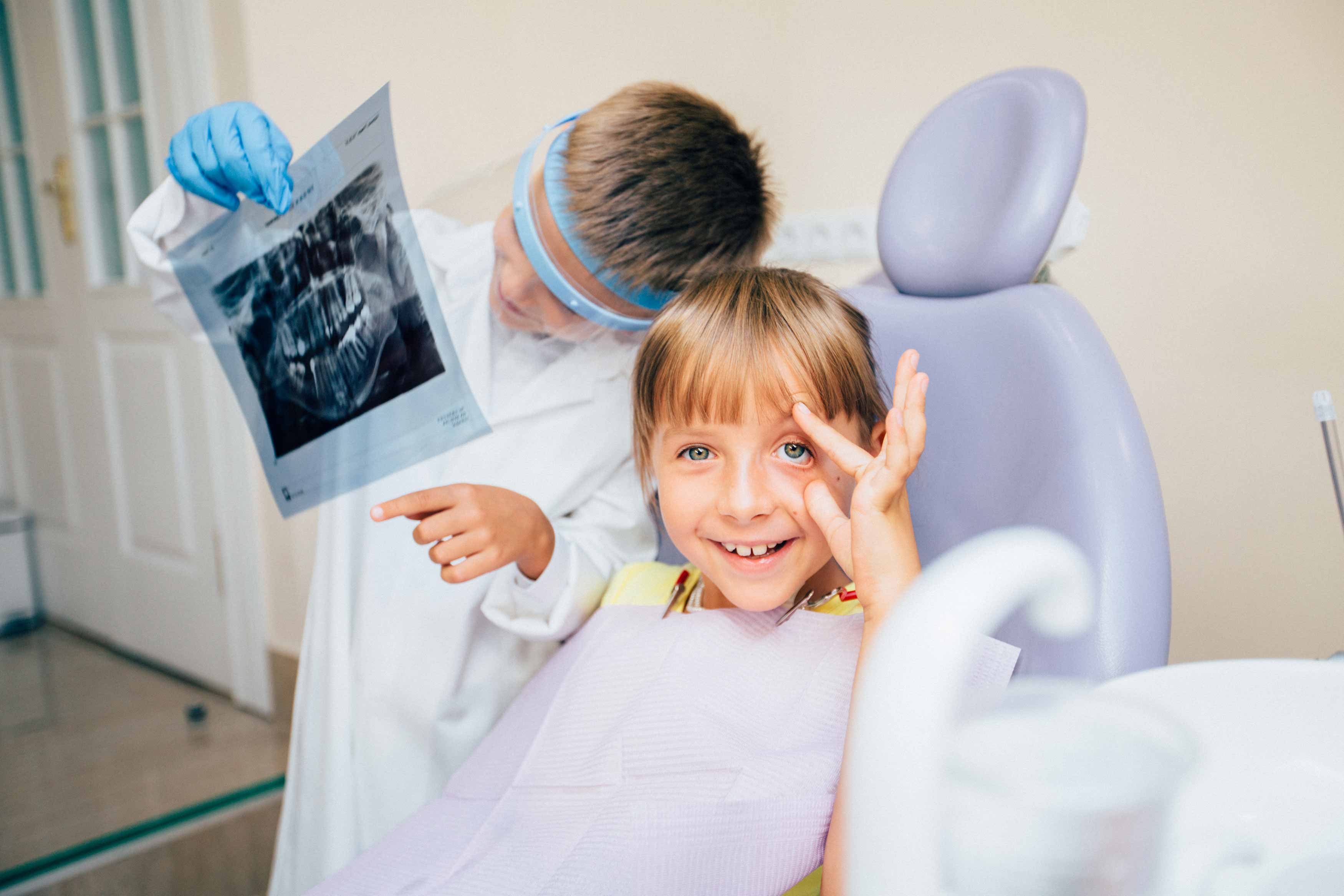 A young girl sitting on the dental chair smiling while a little boy dressed as a dentist showing her an Xray of teeth