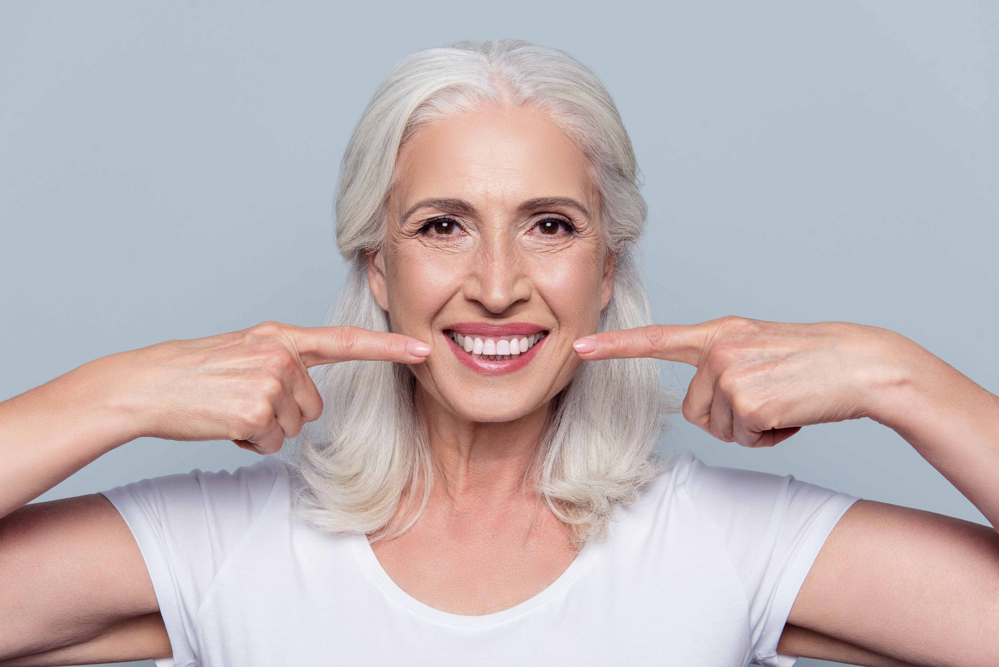 Photo of an elderly female smiling and using two fingers pointing towards her smile. 
