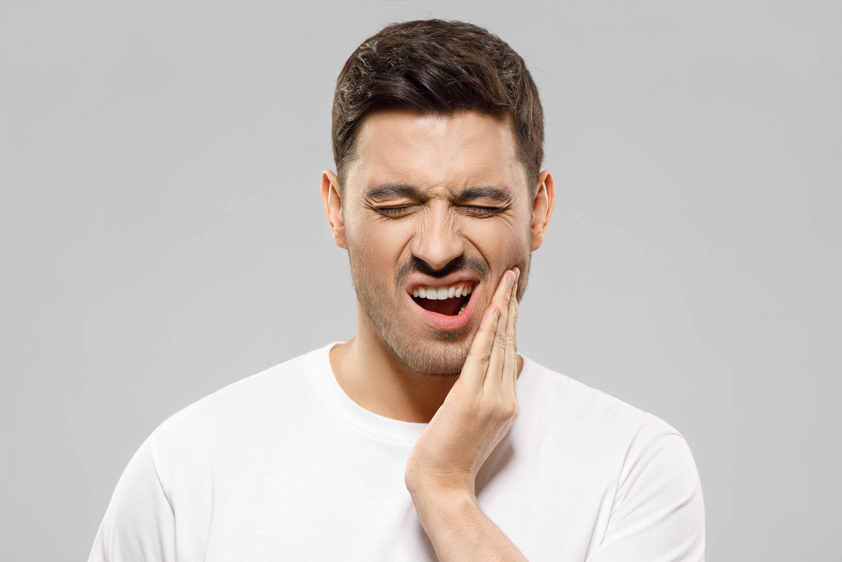 Image of a male in a white t-shirt against a light gray background, touching his left cheek because of a toothache
