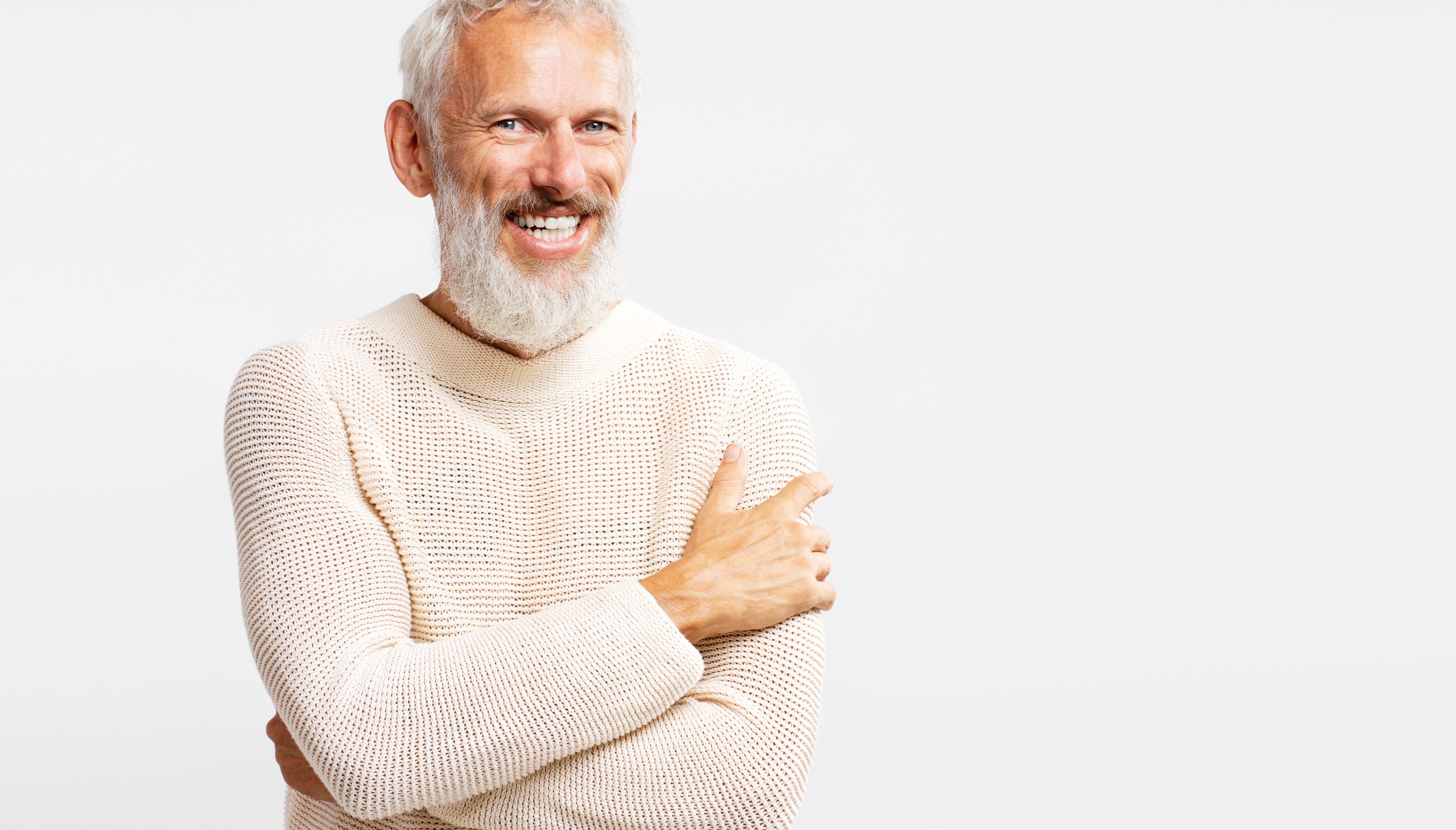 Smiling bearded man wearing stylish warm sweater hugging himself, looking at camera isolated on gray background. Concept of showing off his implant-teeth