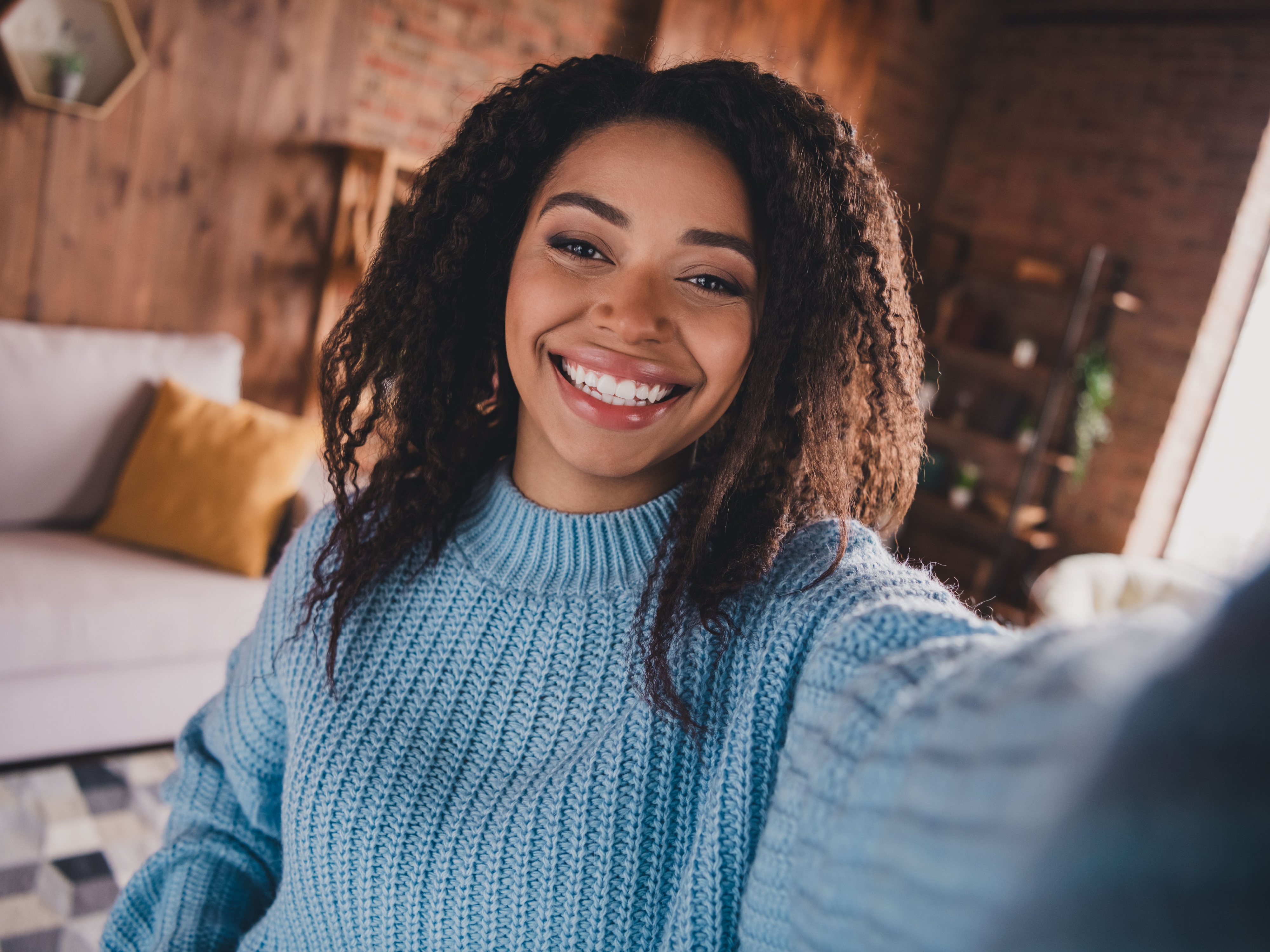 Charming young woman with afro hair smiling in a cozy living room during daylight, wearing a stylish knitted sweater after getting a clean by dentists of hawthorn