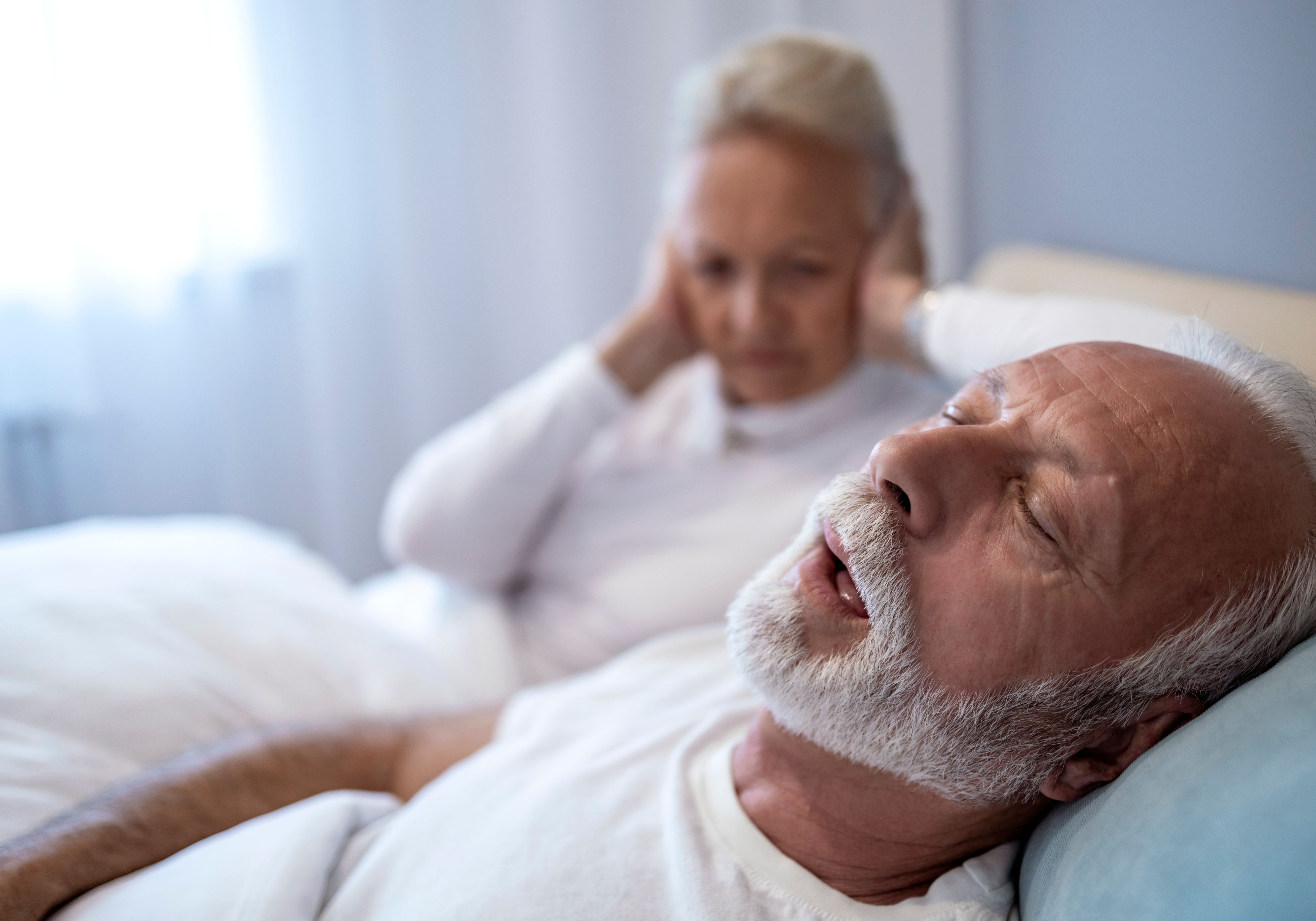 An elderly couple is in bed, with one partner trying to sleep while the other snores loudly. The room is softly lit, creating a peaceful yet challenging night for the couple.